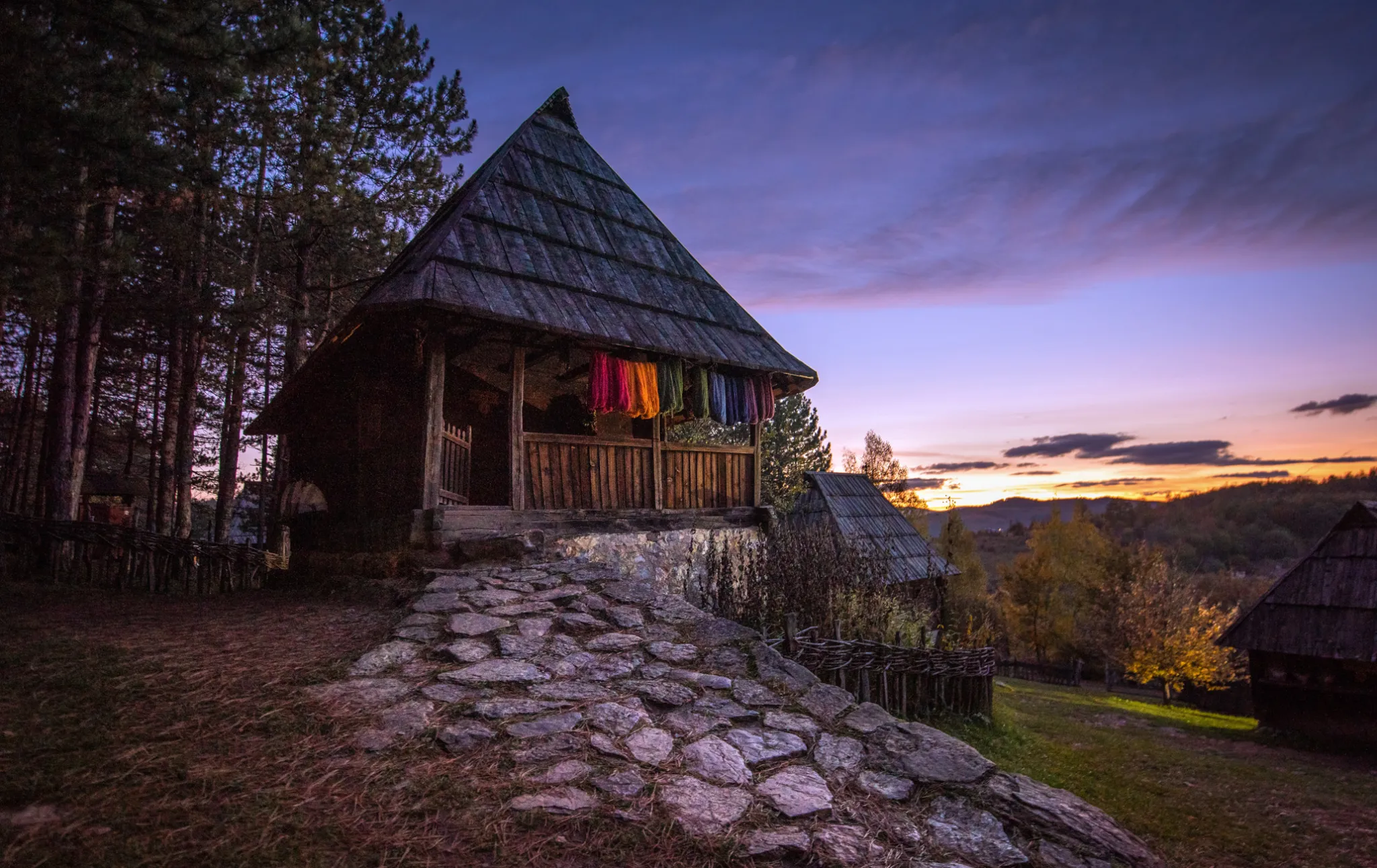 A log house of the Sirogojno open-air museum at dusk with the hills of Zlatibor behind