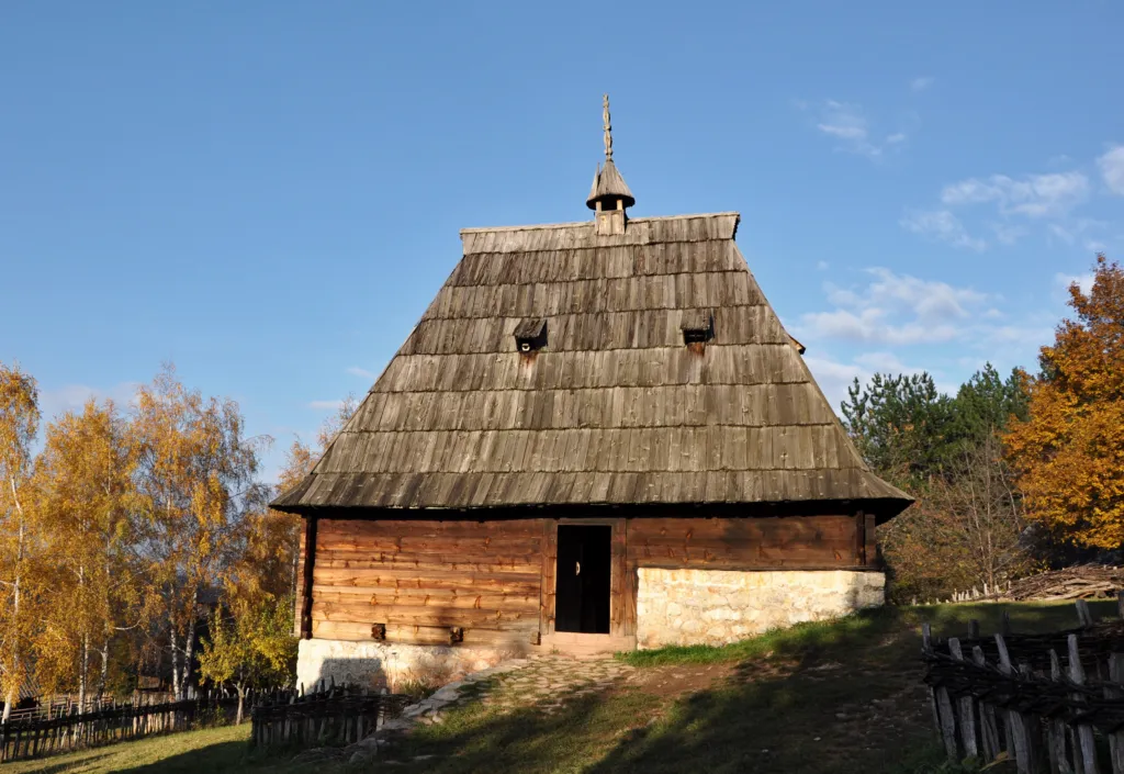 A steep shingle-roofed log house among golden autumn trees in the Sirogojno open-air museum on Zlatibor