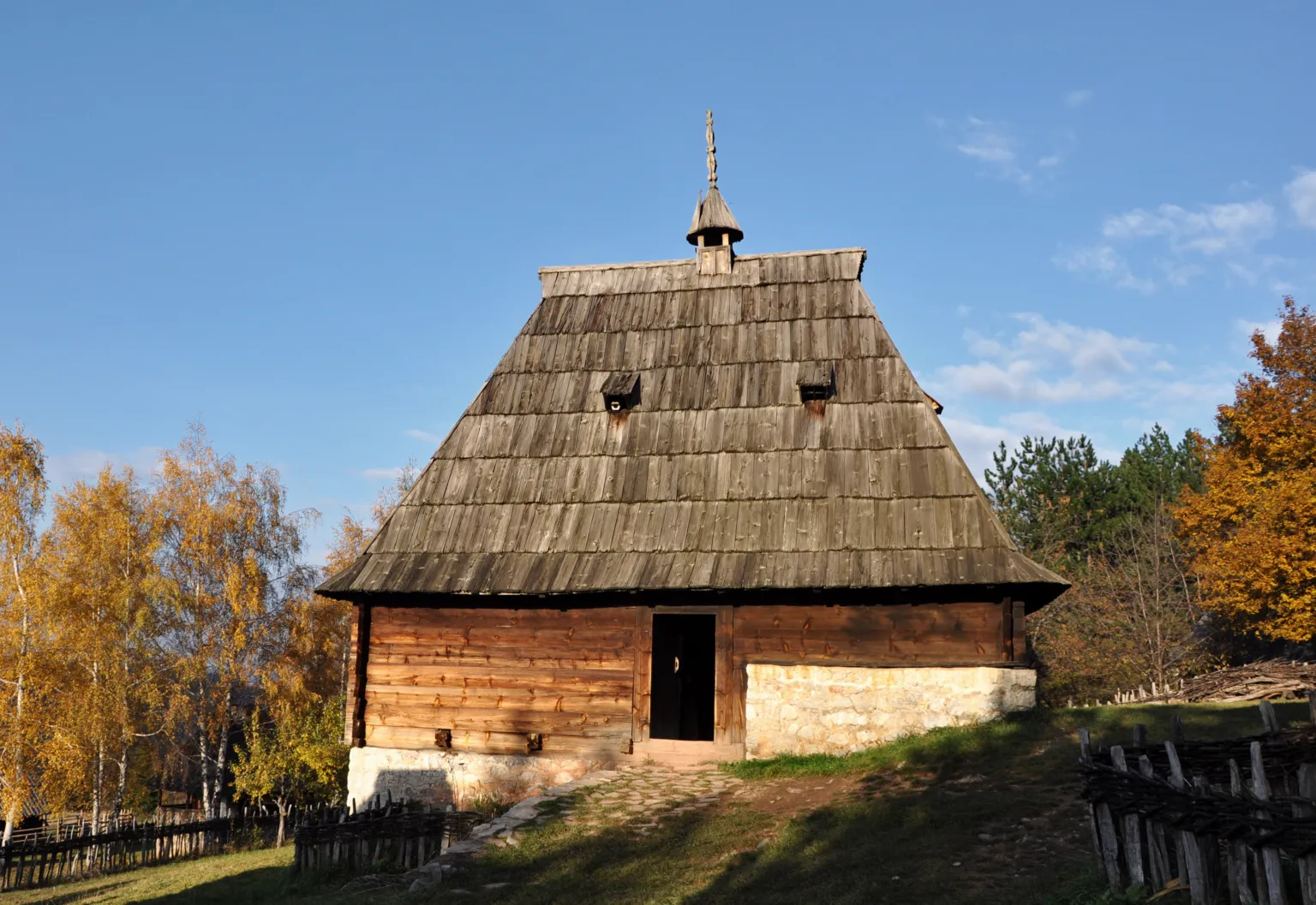 A steep shingle-roofed log house among golden autumn trees in the Sirogojno open-air museum on Zlatibor