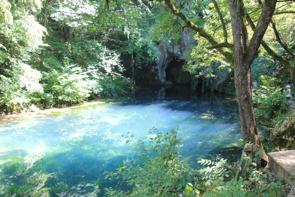 The clear blue pool of Krupaj Spring below a cave mouth in the woods